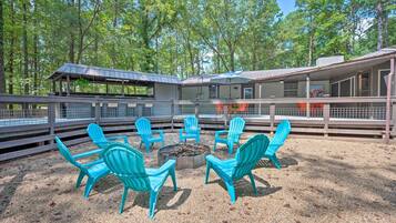 Un espace extérieur aménagé avec des sièges, protégé par un toit, doté de chaises turquoise et d’un foyer, le tout entouré d’une clôture en bois et d’arbres.