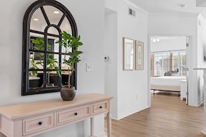 Un pasillo con un espejo arqueado grande, una planta en maceta, una consola de mesa de color claro y una cama al fondo.