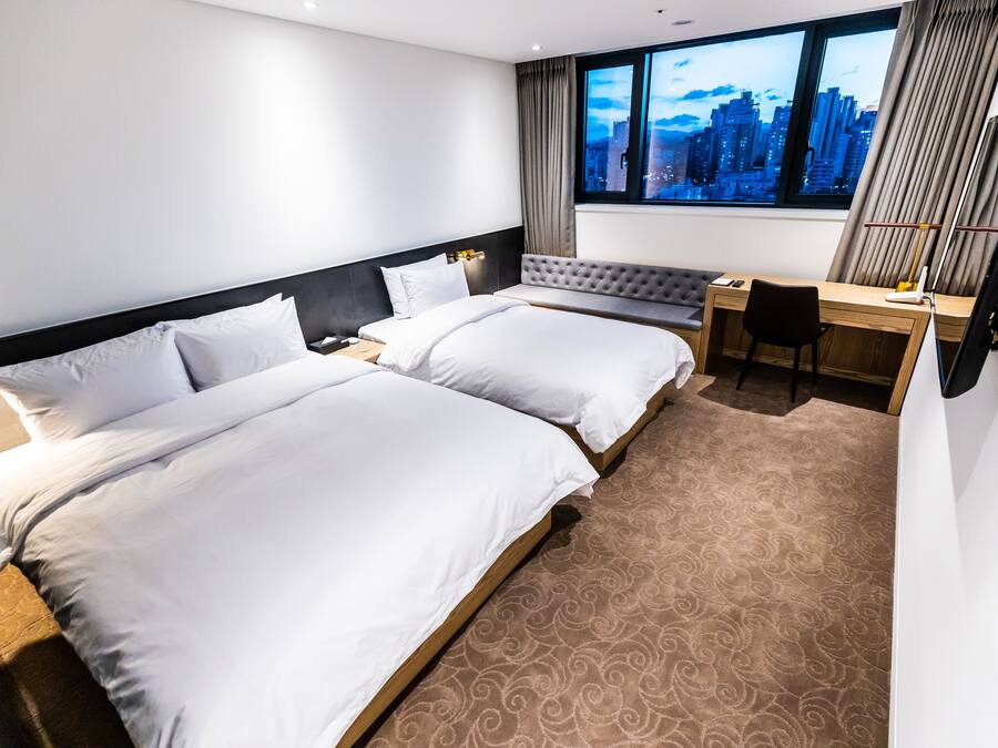 A hotel room with two beds, a desk, and a view of a cityscape through large windows.