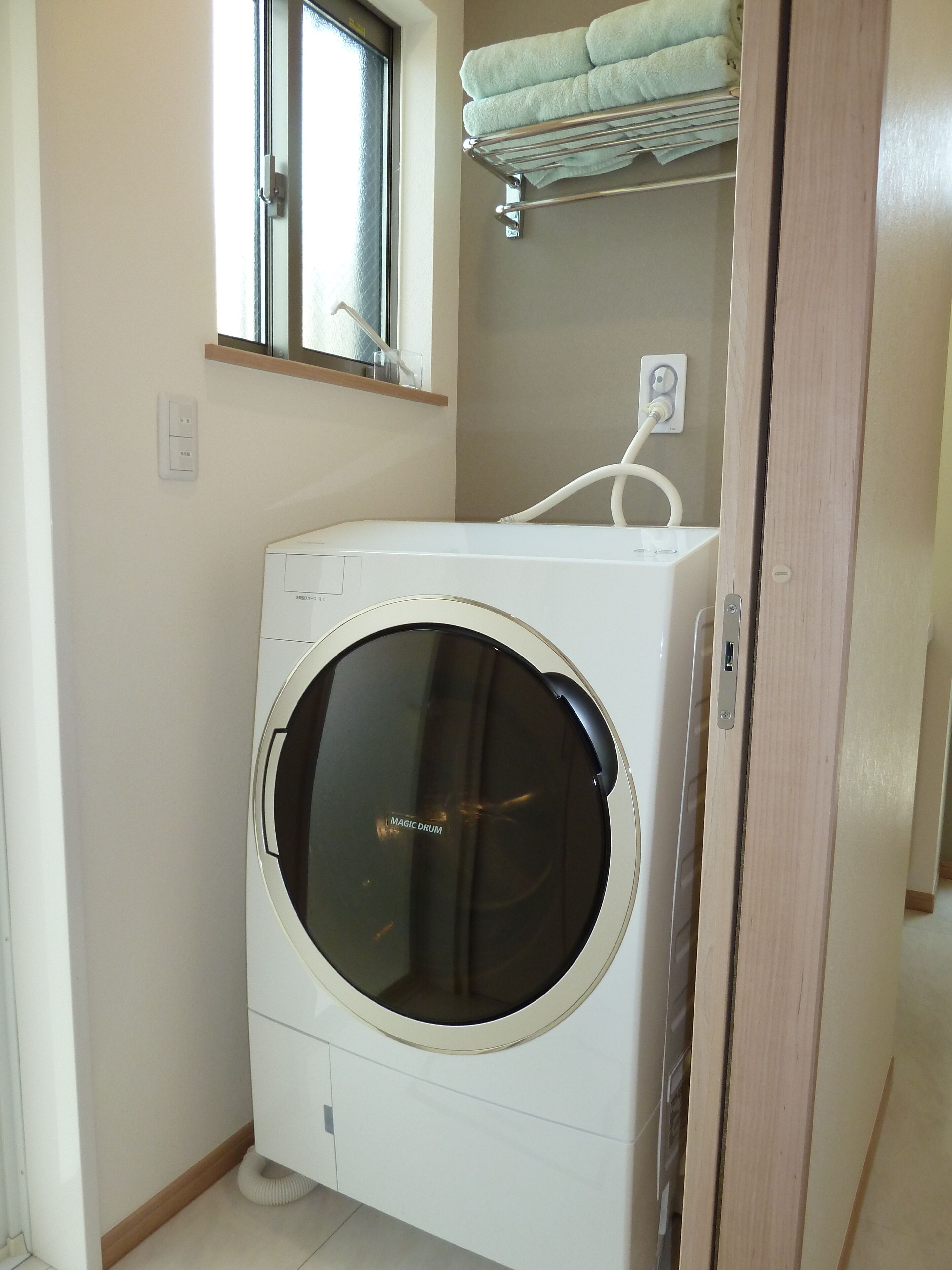 A compact, white front-loading washing machine in a small laundry room with a window above it and a towel rack on the wall.