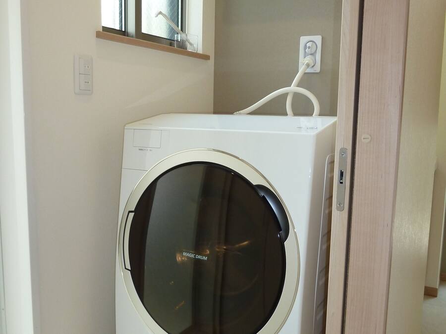 A compact, white front-loading washing machine in a small laundry room with a window above it and a towel rack on the wall.
