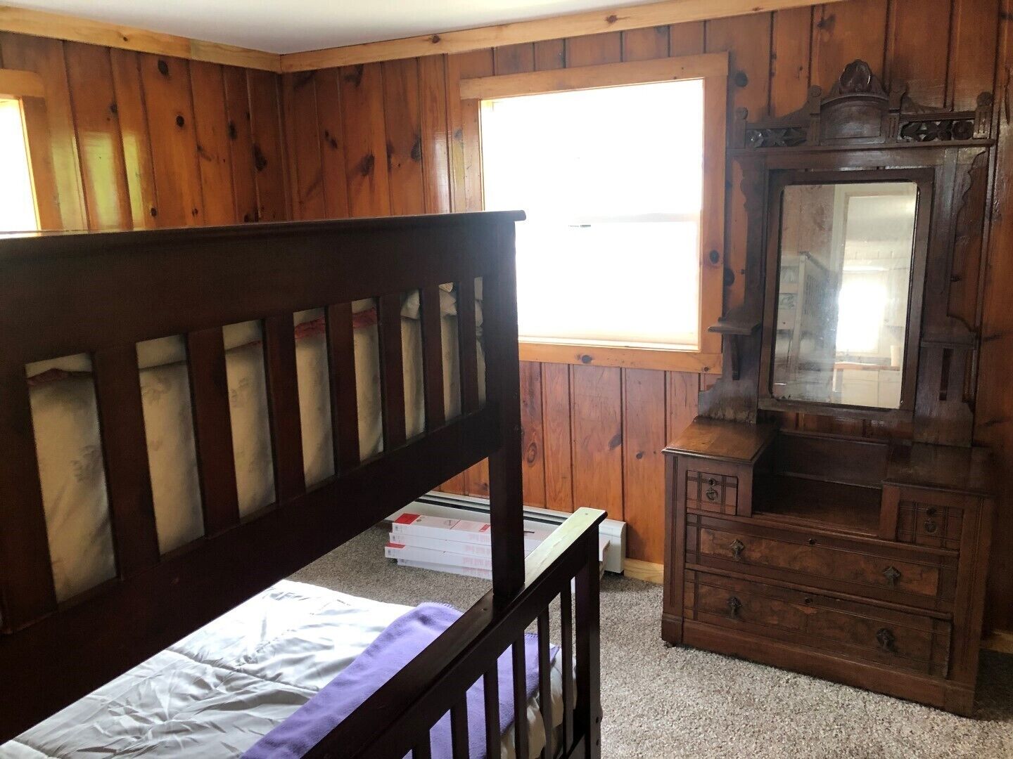 Ein Zimmer mit Holzwänden, einem Bett mit Kopfstütze, einem Schminktisch und einem Fenster, das für natürliches Licht sorgt.