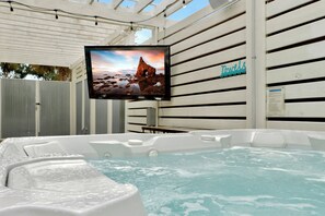 Un jacuzzi con una pantalla de televisión mostrando una escena de atardecer, un letrero que dice 'Toallas' y una estructura de pérgola blanca.