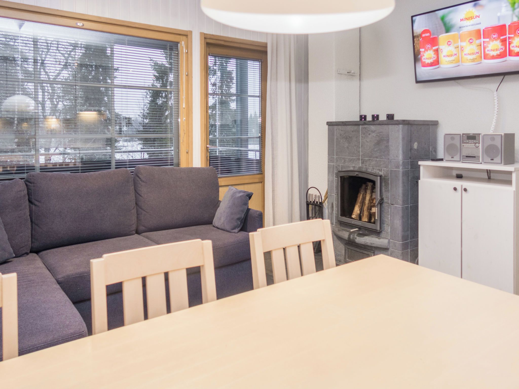 A modern living room with a grey sofa, wooden chairs, a dining table, a fireplace, and a TV mounted on the wall.