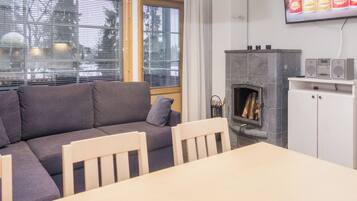 A modern living room with a grey sofa, wooden chairs, a dining table, a fireplace, and a TV mounted on the wall.