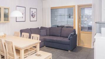 A modern living room with a grey sofa, wooden dining table, and chairs. There are framed pictures on the wall, a window with blinds, and a door.