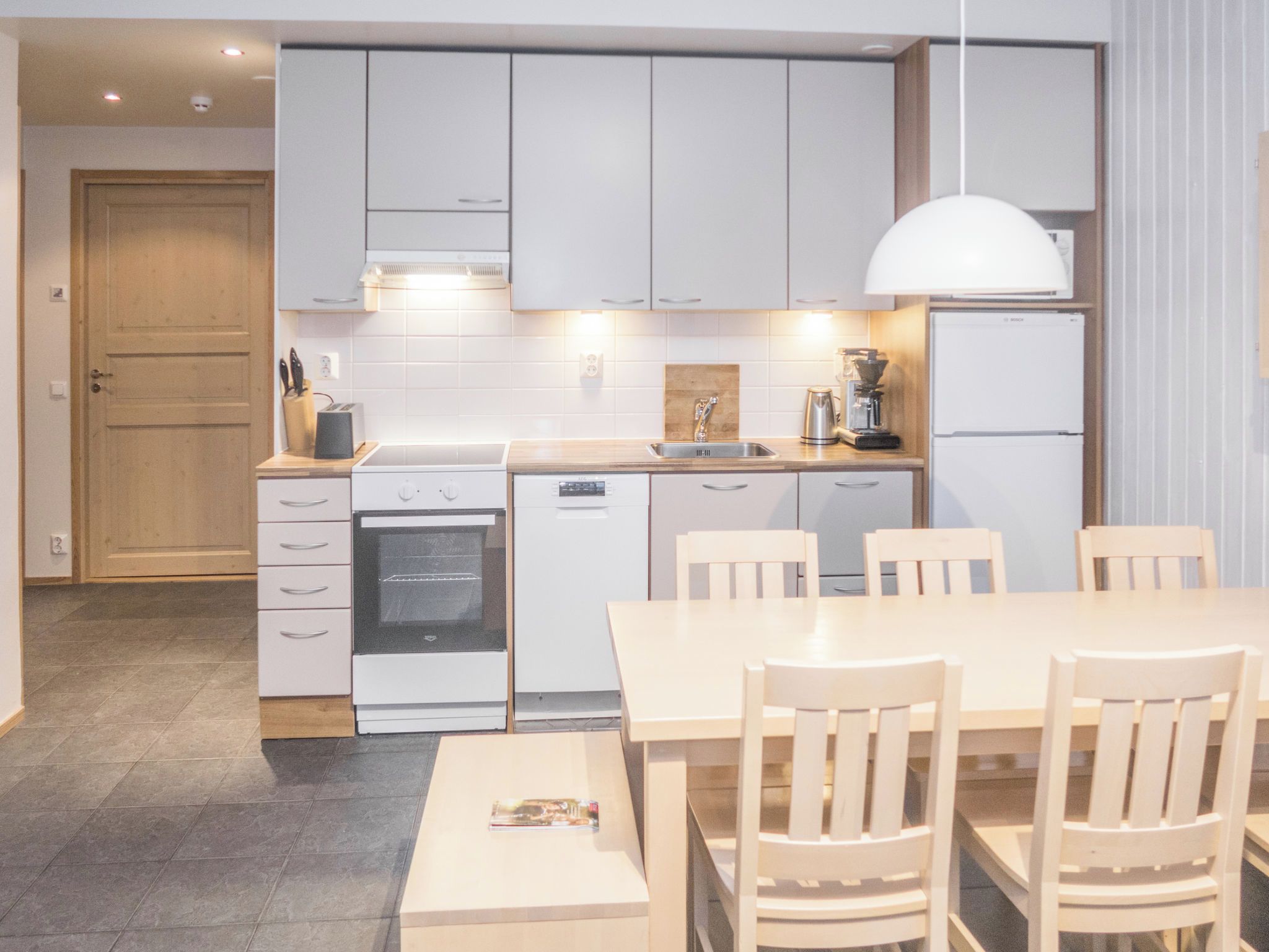 A modern kitchen with white cabinets, a built-in oven, a coffee machine, and a dining table with chairs.