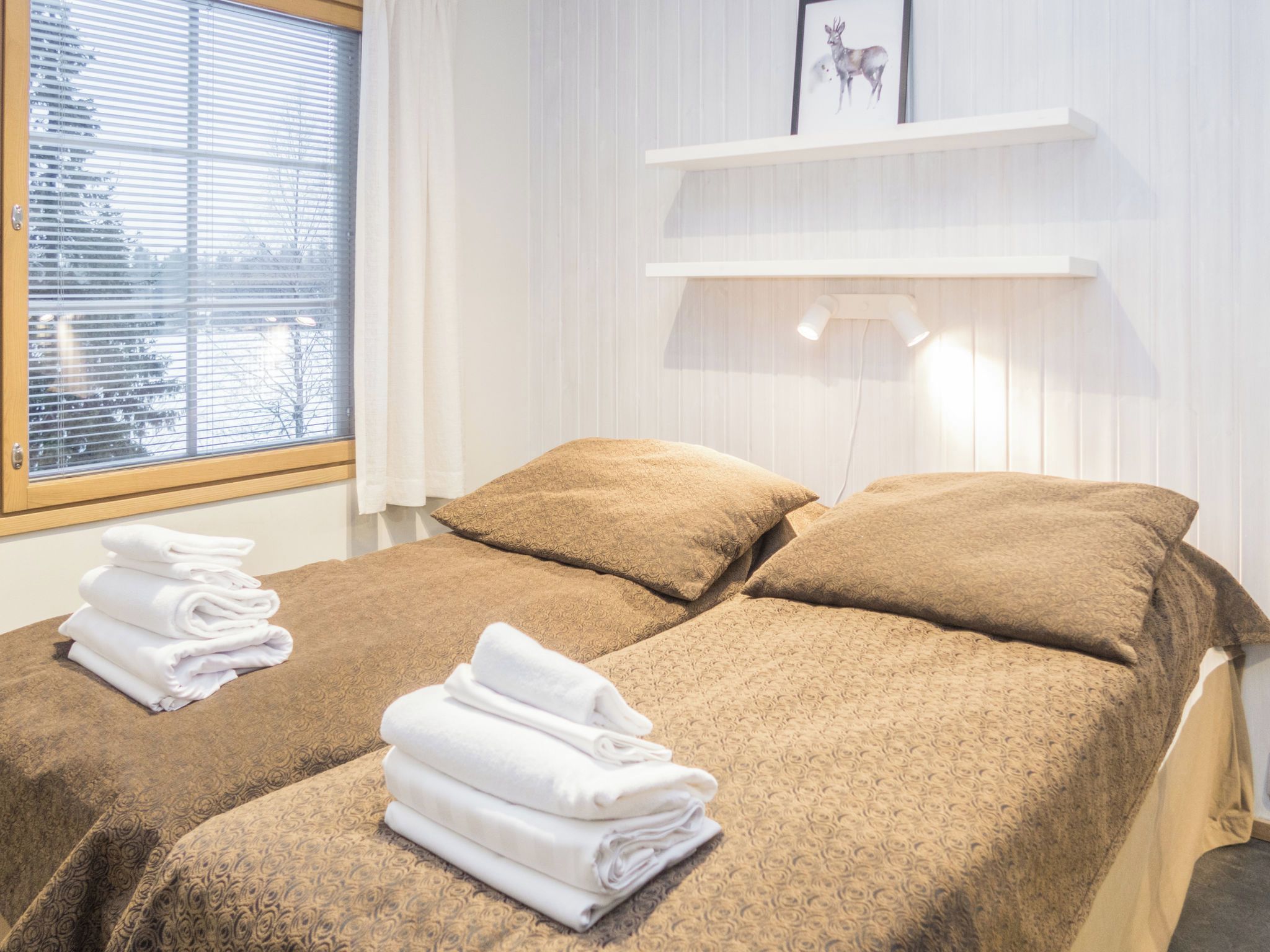A hotel room with two beds, each with a brown bedspread and white towels. There is a window with blinds, a white wall with a shelf, and a picture frame.
