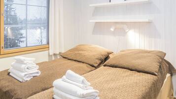 A hotel room with two beds, each with a brown bedspread and white towels. There is a window with blinds, a white wall with a shelf, and a picture frame.