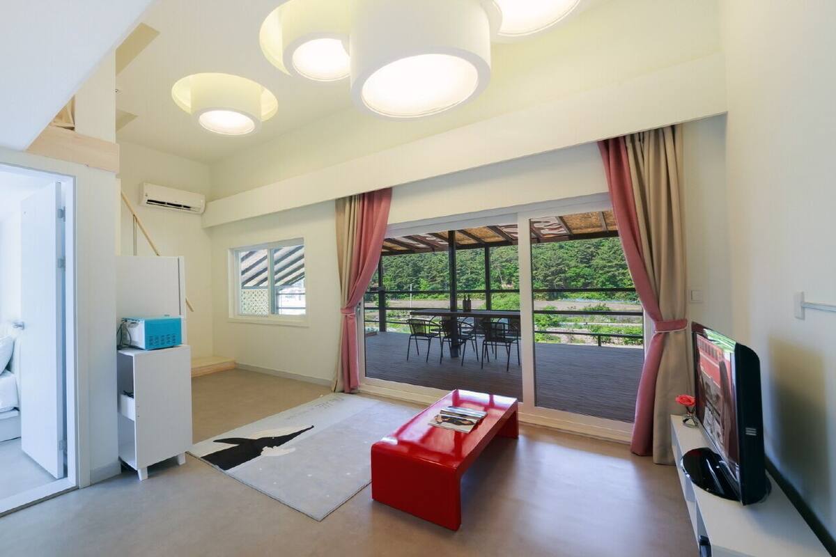 Une chambre moderne équipée d’un lit, d’un bureau, d’un banc rouge, d’une télévision et de grandes fenêtres donnant sur un paysage verdoyant.