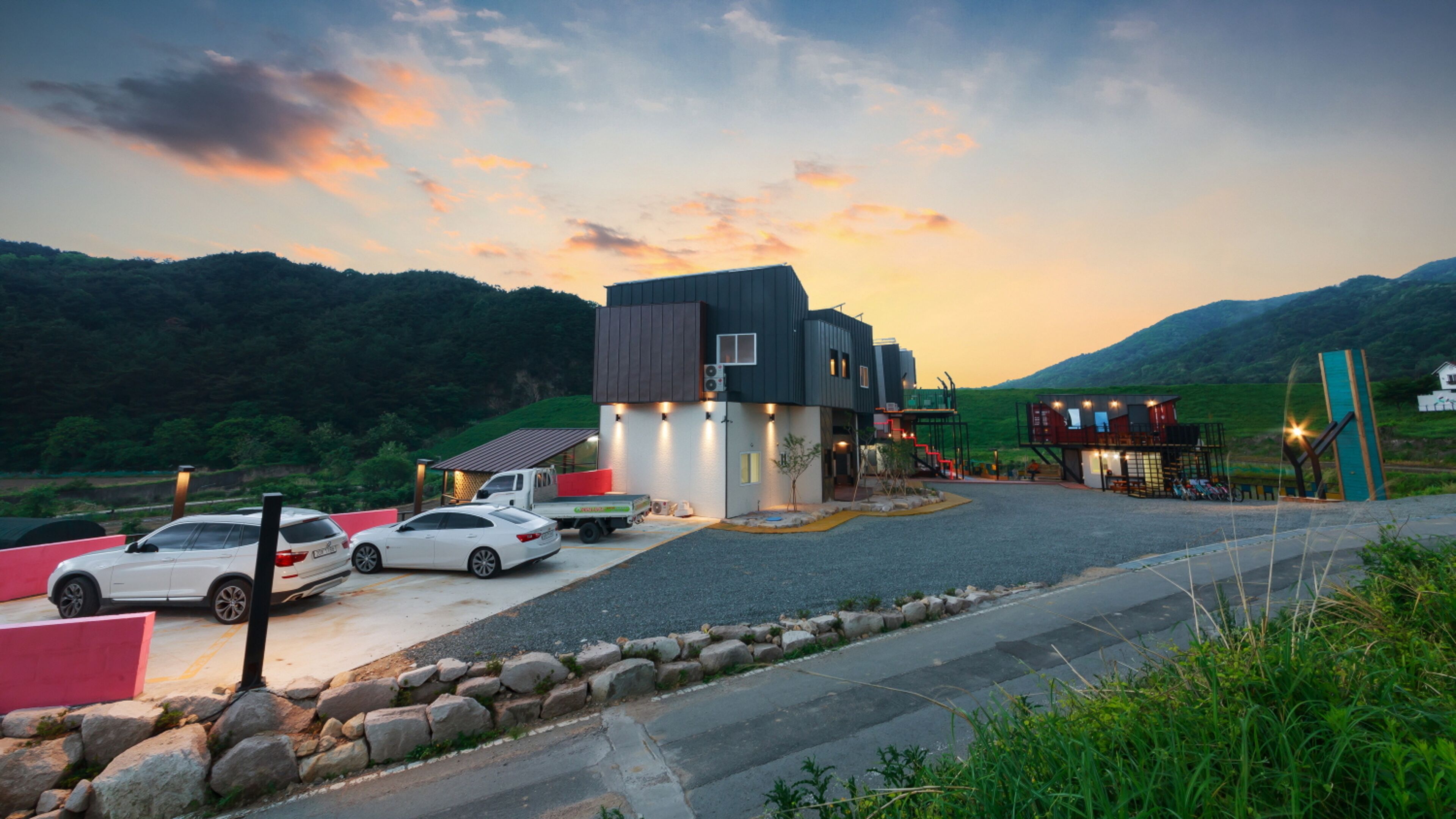 A modern building with a flat roof and a large entrance. There are two white cars parked in front.