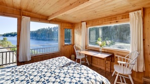 Une cabane en bois comprenant un lit, une table, deux chaises et de grandes fenêtres donnant sur un lac et une forêt.