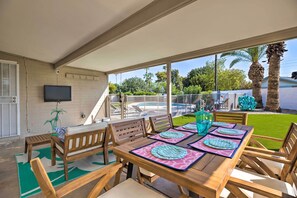 Eine überdachte Terrasse mit Holzmöbeln, einem an der Wand befestigten Flachbildfernseher und Blick auf den Garten.