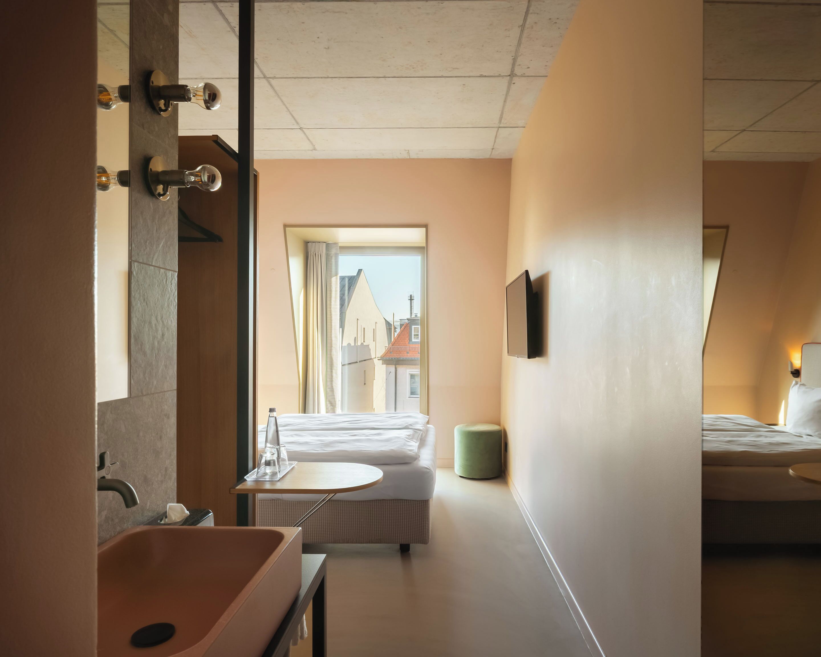 A modern hotel room with a large mirror, a sink, a bedside table, a bed, and a view of the city through the window.