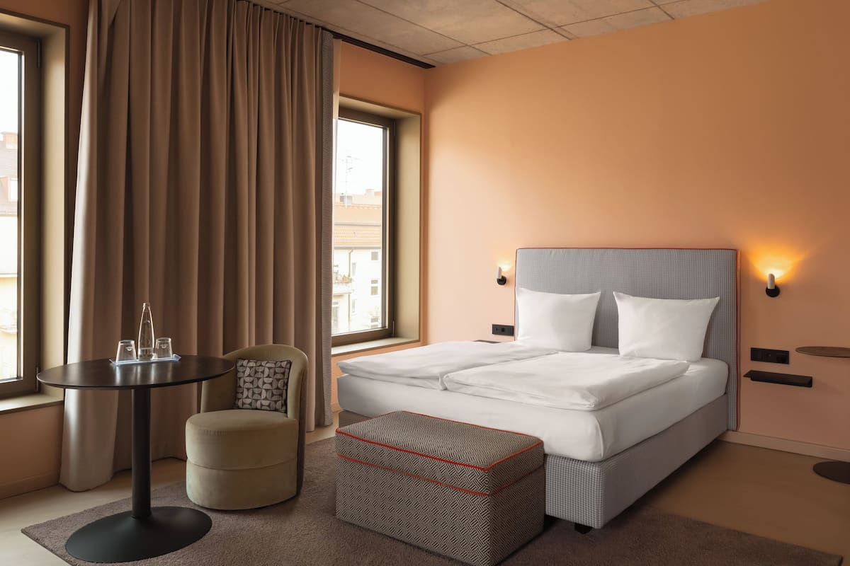 A hotel room with a bed, a small table with glasses, a chair, and a window with curtains.