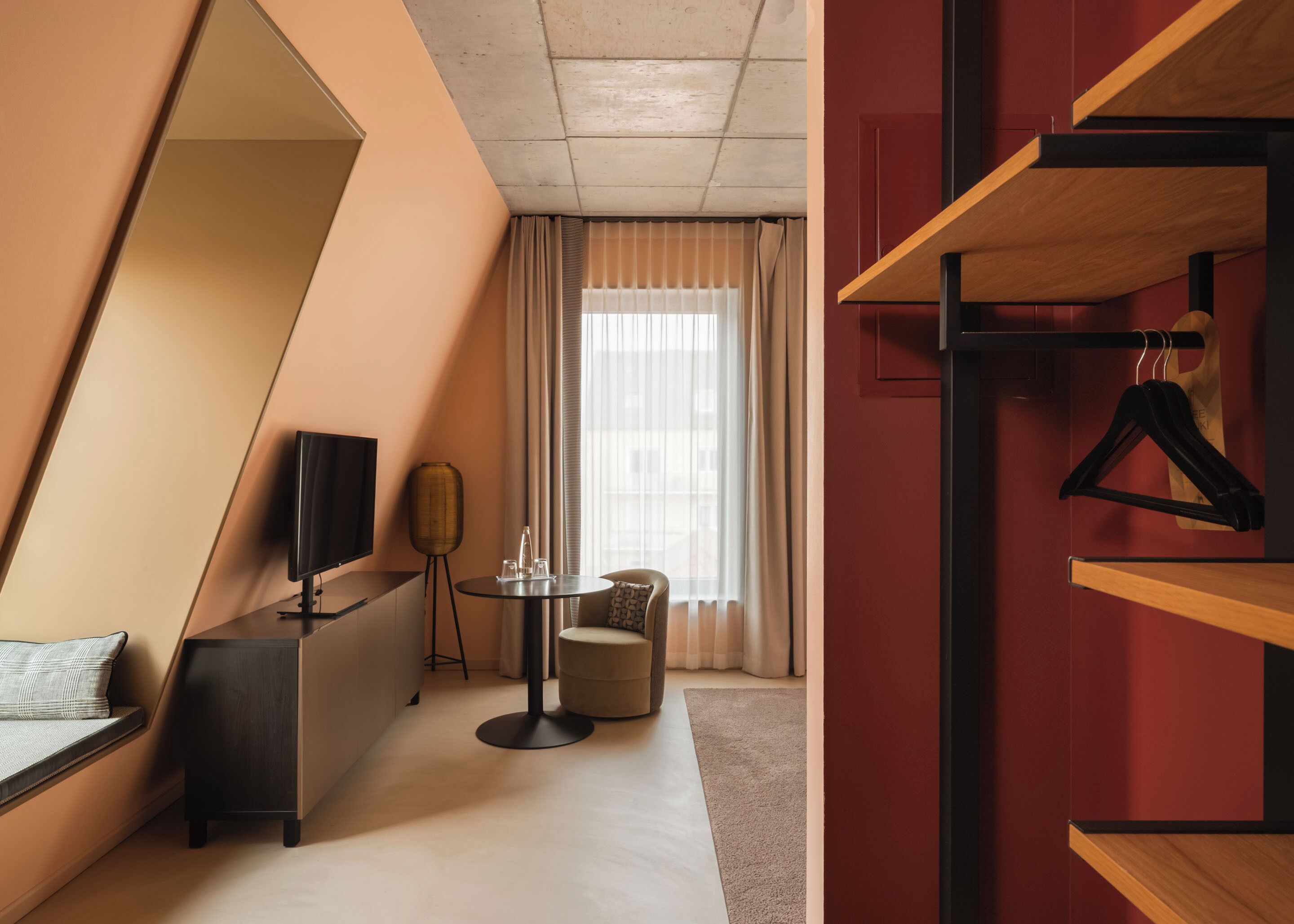 A modern hotel room with a large mirror, a TV, a round table, a sofa, and a wardrobe with hangers.