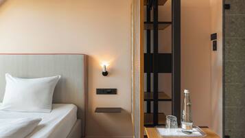 Une chambre d’hôtel avec un lit bien fait, une table de chevet sur laquelle se trouvent une bouteille et un verre, et une lampe fixée au mur.
