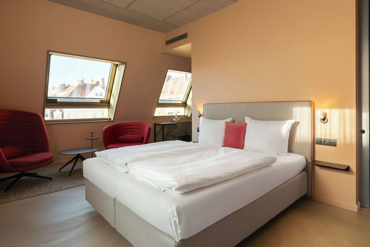 A hotel room with a large bed, two red chairs, a small table, and two windows with city views.
