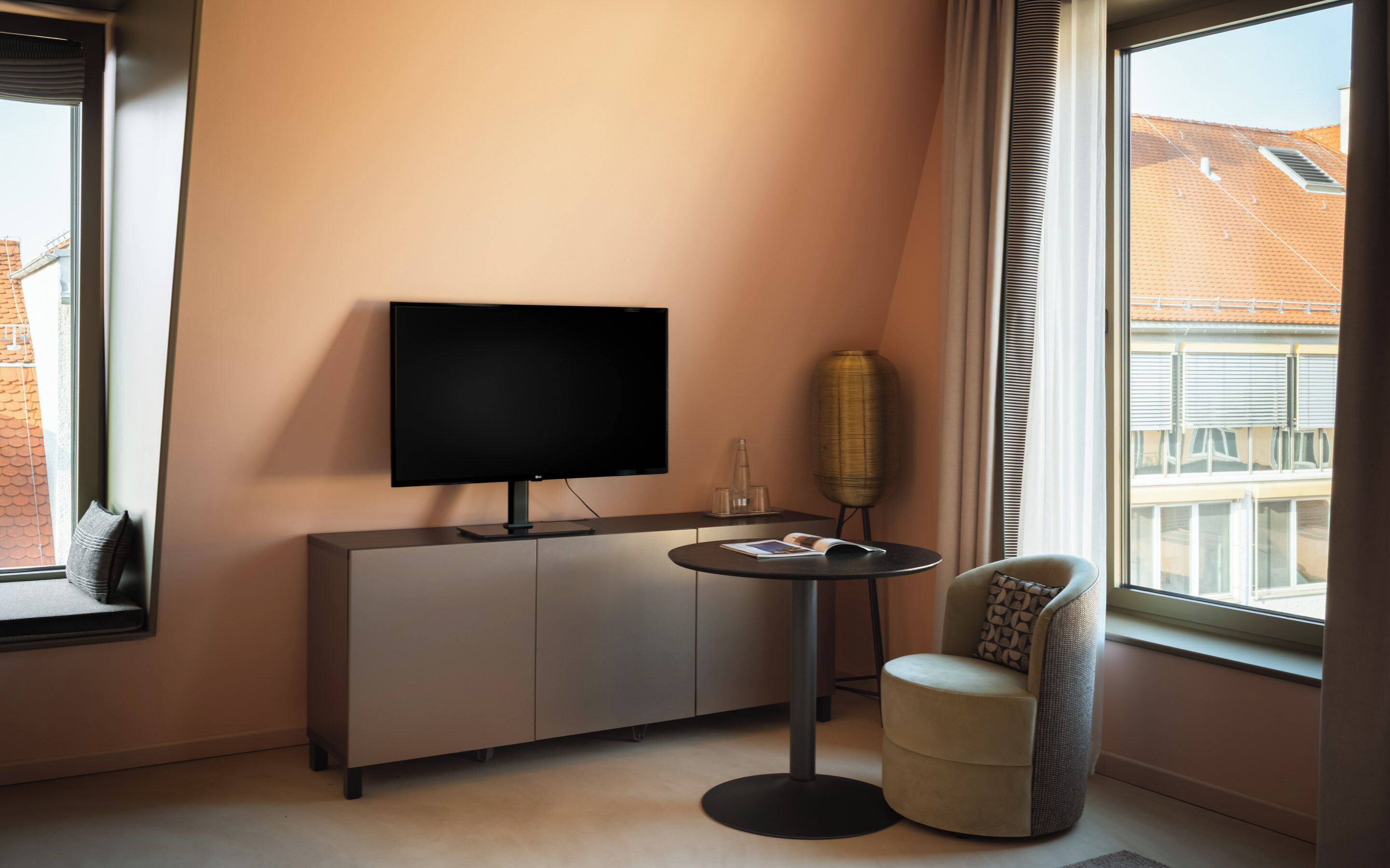 A modern living room with a flat-screen TV, a round table, a chair, and a large window with a view of a building.