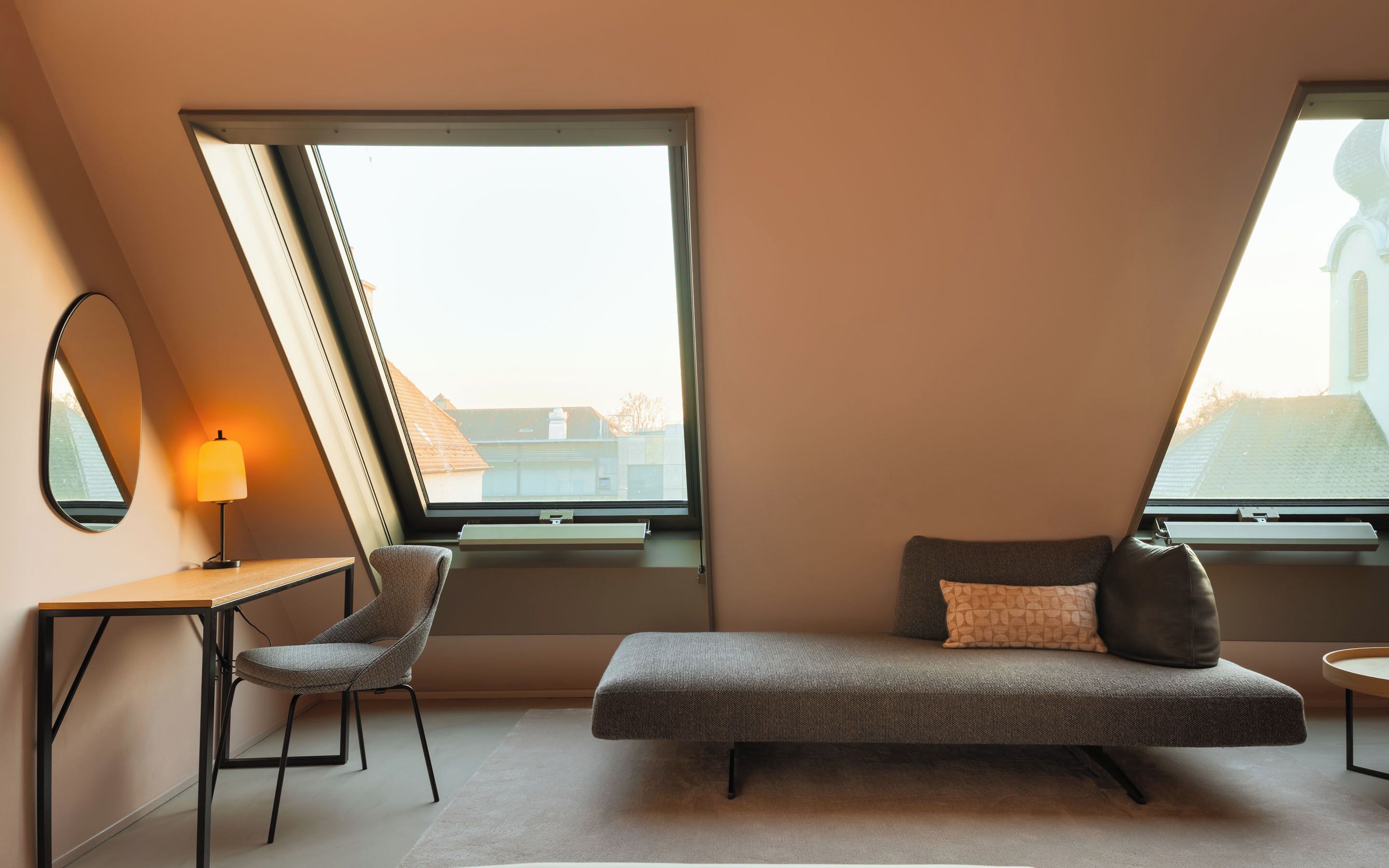 A modern loft bedroom with a sloped ceiling, a desk with a chair, a sofa with cushions, and large windows offering a view of buildings outside.
