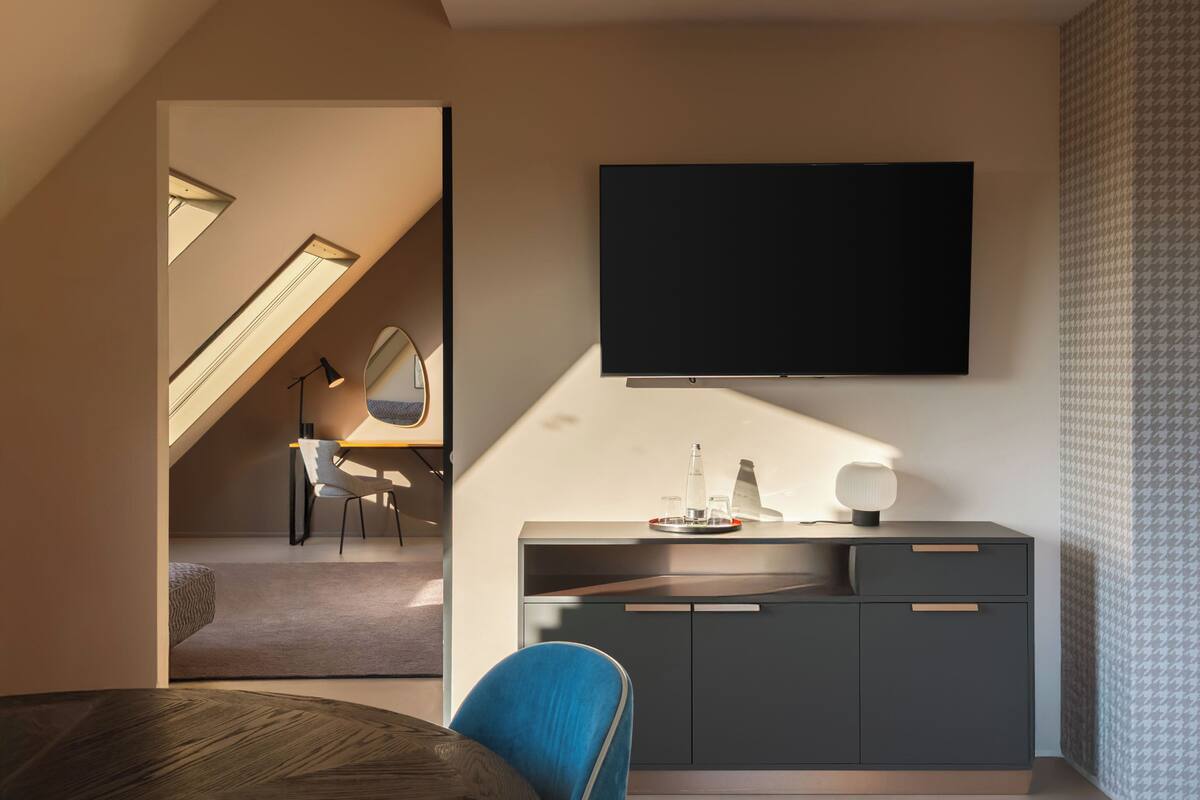 A modern living room with a large flat-screen TV mounted on the wall, a wooden dining table, a blue chair, and a staircase leading to an upper level.