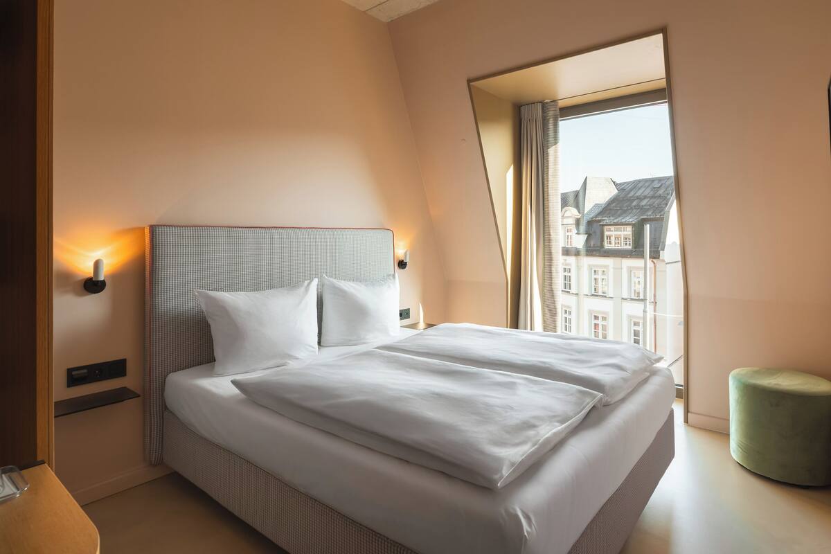 A neatly made bed in a hotel room with a view of buildings through a window.