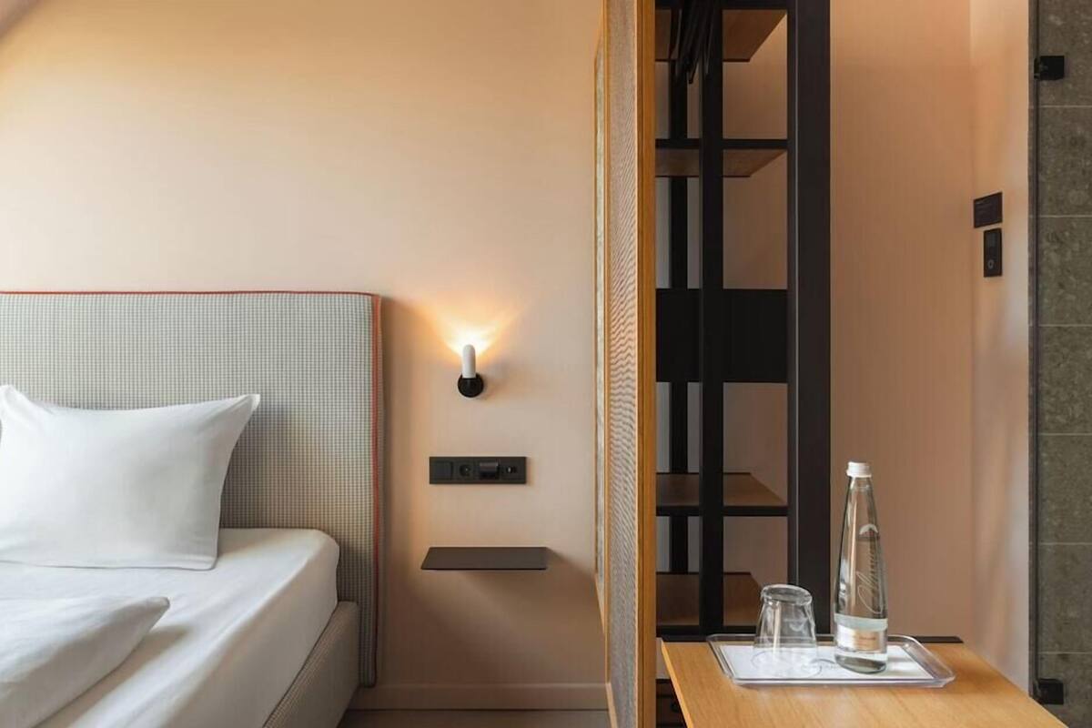 A hotel room with a bed, a nightstand, a wall-mounted light, a bottle, and a tray with glasses.