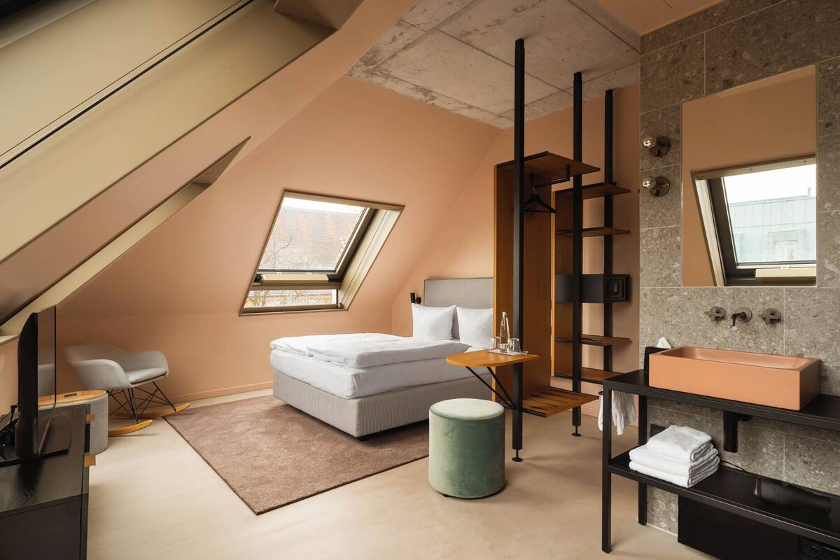A modern bedroom with a skylight, a bed, a bedside table, a green ottoman, a vanity with a mirror, and a bathroom with a sink and towel rack.