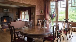 Een klassieke kamer met een open haard, een ronde eettafel met stoelen en grote ramen met gordijnen.