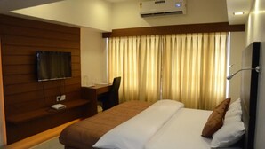 A hotel room with a bed, a desk, a chair, a TV, and an air conditioning unit. - Hotel Yuvraj (Vadodara)