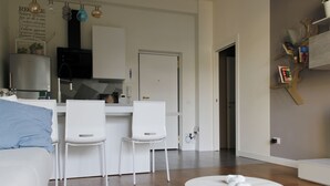 Un salón moderno con un sofá blanco, dos sillones blancos, una mesa de centro redonda y una zona de cocina con un armario blanco y una campana extractora negra.