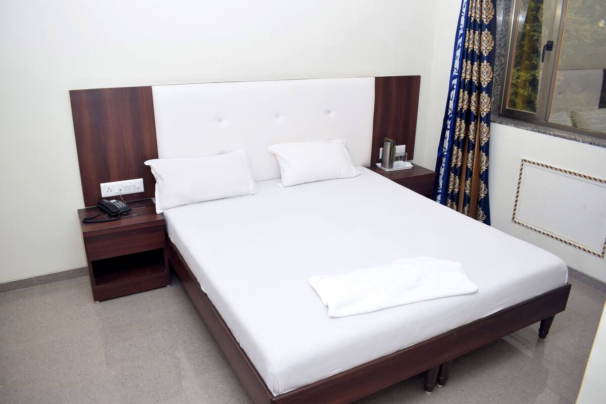 Een net opgemaakt bed met witte lakens, een houten hoofdbord, een nachtkastje met een telefoon, en een raam met gordijnen.