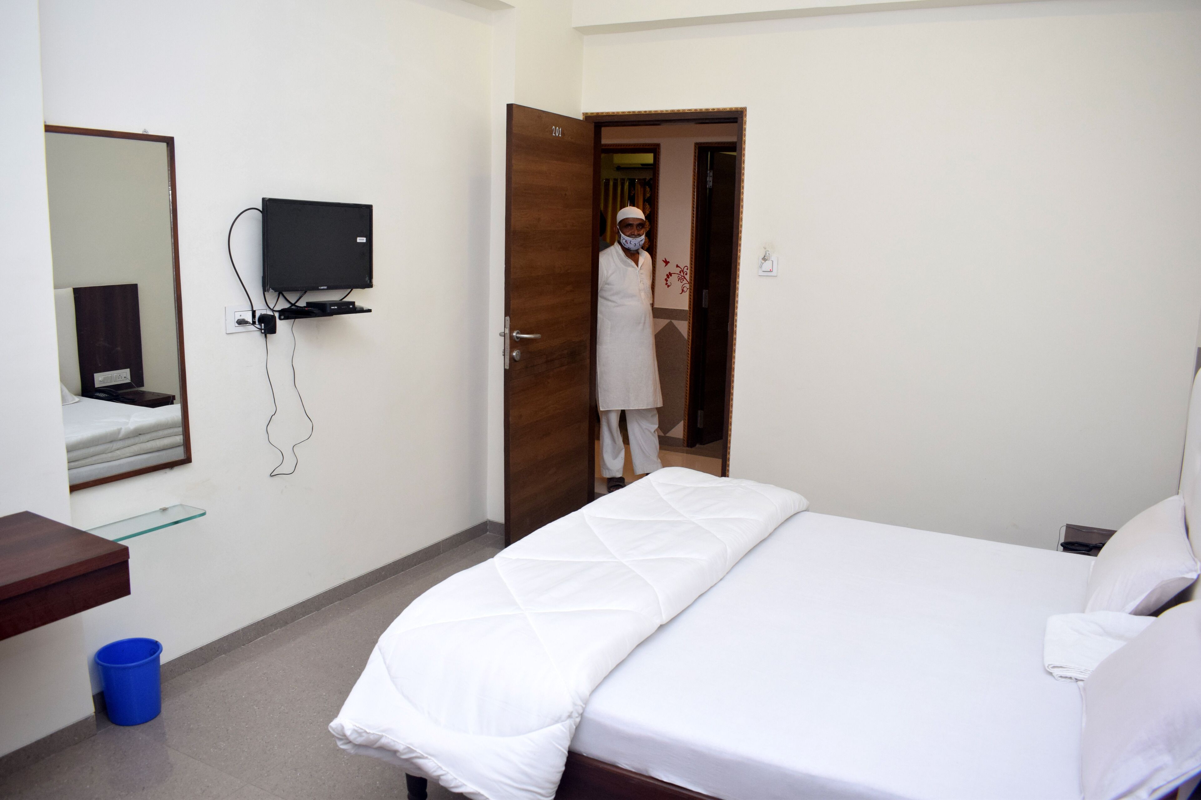 A hotel room with a bed, a TV mounted on the wall, a mirror, and a blue trash bin.