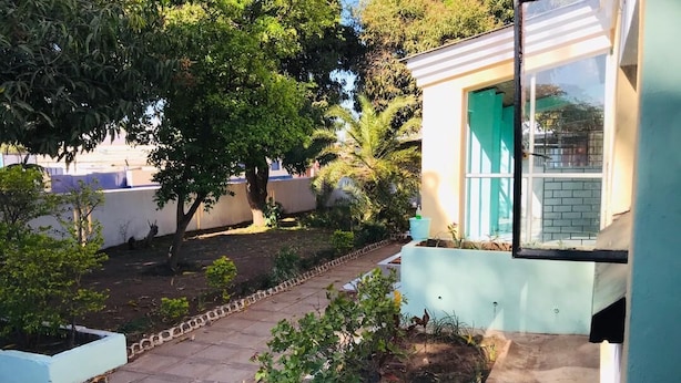Petit edifici amb una porta verda i una finestra. Hi ha un camí pavimentat que condueix a l'edifici, envoltat de plantes i arbres petits.