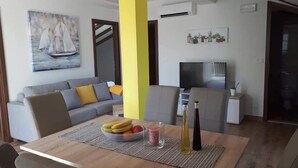 A modern living room with a sofa, a dining table with fruit and drinks, a TV, and a painting of a sailboat. - Apartments Šišević (Dubrovacko Primorje)