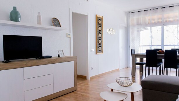 A modern living room with a flat-screen TV, a wooden cabinet, a dining area with a table and chairs, and artwork on the wall.