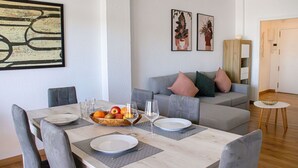 A dining area with a marble table, grey upholstered chairs, and a grey sofa with cushions. There are two framed artworks on the wall, a fruit bowl on the table, and a small round table with a lamp.