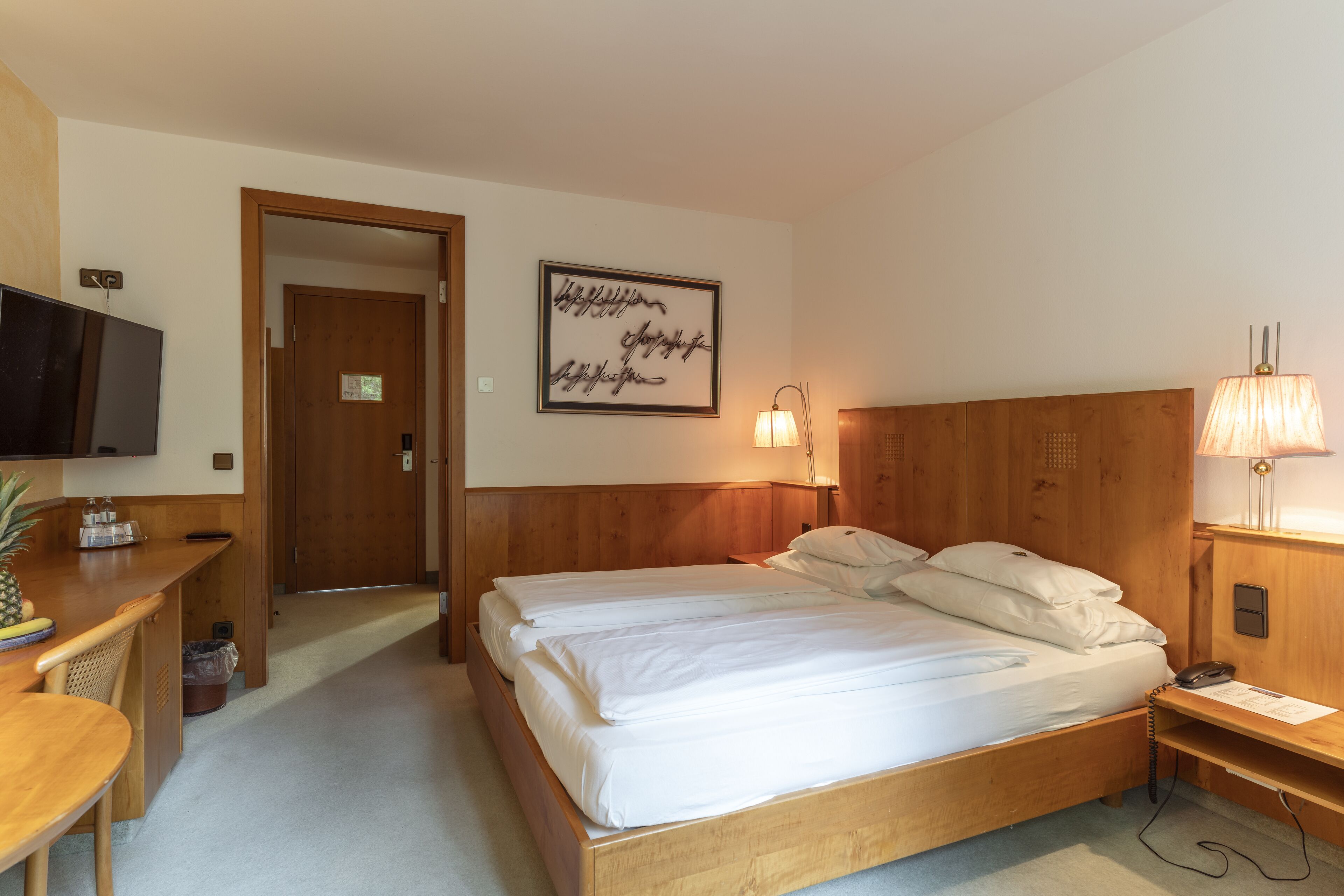 A hotel room with a wooden bed, a desk with a television, a wall-mounted lamp, and a framed quote on the wall.