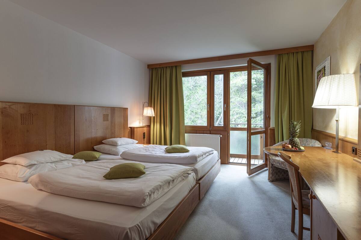 Ein Hotelzimmer mit zwei Betten, einem hölzernen Kopfteil, einem Esstisch mit Stühlen und einem Fenster mit Vorhängen.