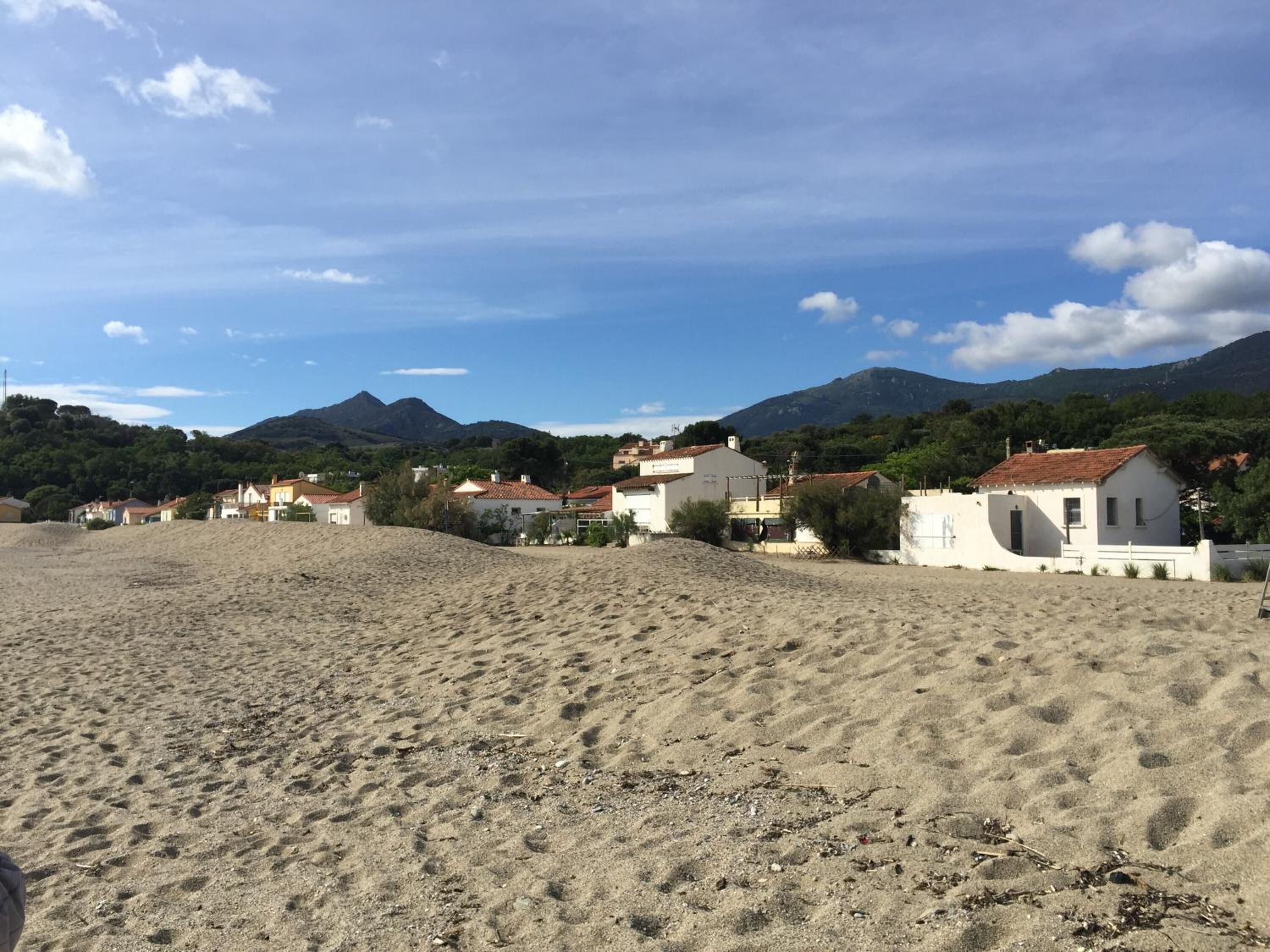 A beach with white buildings, a sandy foreground, and mountains in the background.