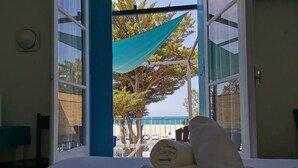 A hotel room with a bed, a window with a view of a beach and palm trees, and a blue awning outside. - Hotel Oasis (Argelès-sur-Mer)