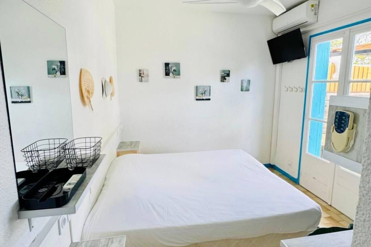 A compact room with a bed, a small table, a chair, a ceiling fan, and a window with blue shutters.