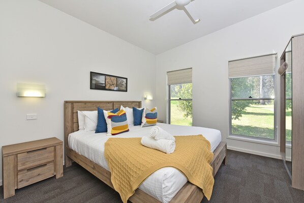 A bedroom with a bed, bedside tables, a wall-mounted light, a window with blinds, and a view of greenery outside. - Ironbark Hill Villa 5 - Vermentino (Pokolbin)