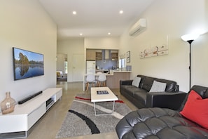 A modern living room with a flat-screen TV, a sofa, a coffee table, and a dining area with a kitchen in the background.