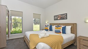A bedroom with a large bed, a wooden headboard, a window with blinds, a bedside table, and a wall-mounted picture.