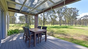A covered outdoor seating area with a table and chairs, overlooking a grassy field with trees.