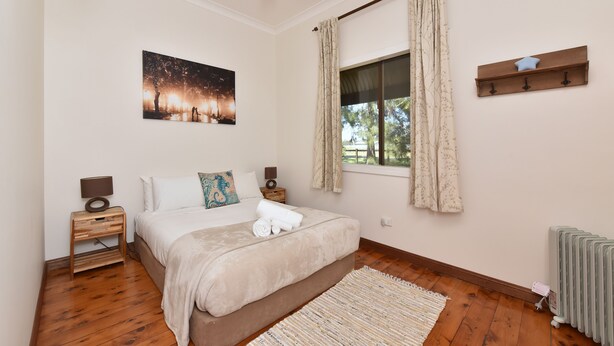 A bedroom with a bed, nightstands, a window with curtains, a radiator, and a wall-mounted shelf.