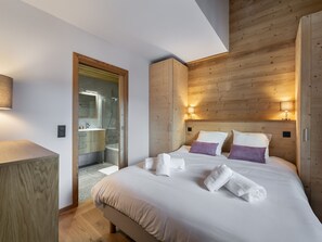 A modern hotel room with a wooden headboard, a large bed with white and purple pillows, a bedside table, and a bathroom visible through an open door.