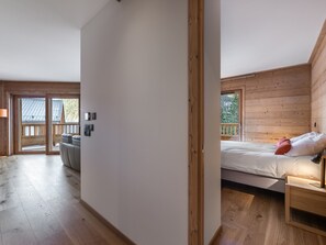 Quarto com cama, mesa de cabeceira, piso de madeira e vista para o exterior.