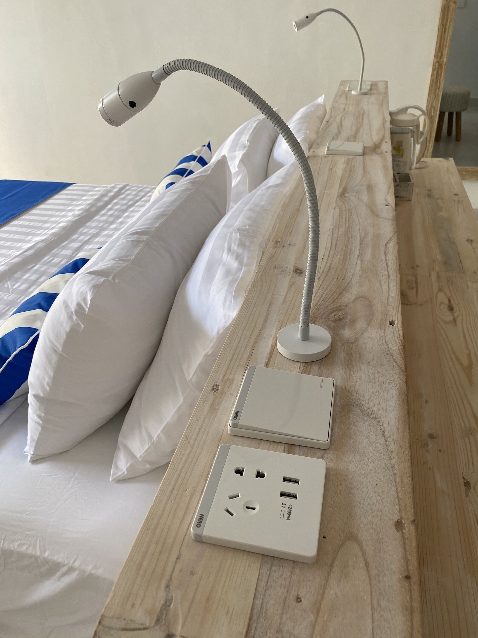 A neatly made bed with white and blue bedding, a wooden bedside table with a lamp and two power outlets.